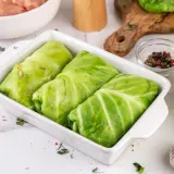 Gotowe do pieczenia gołąbki z młodej kapusty w naczyniu żaroodpornym, obok składniki: mięso mielone, czosnek, przyprawy.