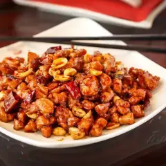 Kurczak King Kong, danie z kawałkami kurczaka w sosie, orzeszkami ziemnymi i papryczkami chili, podane na białym talerzu z pałeczkami.