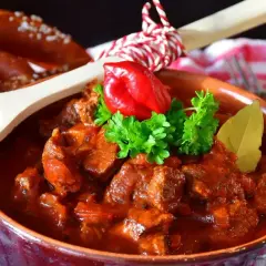 Pyszny gulasz wołowy w glinianej misie, udekorowany papryczką chili i natką pietruszki. Obok leży precel i drewniana łyżka z pieprzem. Idealny przepis na gulasz!