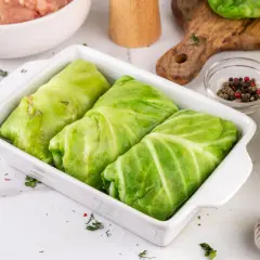 Gotowe do pieczenia gołąbki z młodej kapusty w naczyniu żaroodpornym, obok składniki: mięso mielone, czosnek, przyprawy.