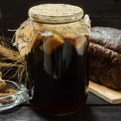 Słoik z ciemnym płynem, chleb żytni i kłosy zbóż – wszystko gotowe do przygotowania domowego kwasu chlebowego.