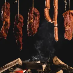 Wędzone mięsa i kiełbasy wiszą nad ogniskiem. Dym unosi się, sugerując, jakie drewno do wędzenia kiełbasy nada najlepszy aromat.