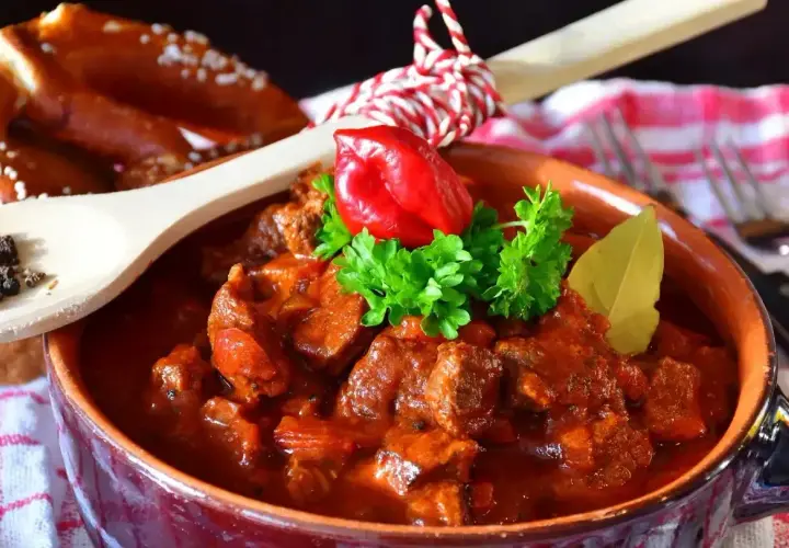 Pyszny gulasz wołowy w glinianej misie, udekorowany papryczką chili i natką pietruszki. Obok leży precel i drewniana łyżka z pieprzem. Idealny przepis na gulasz!