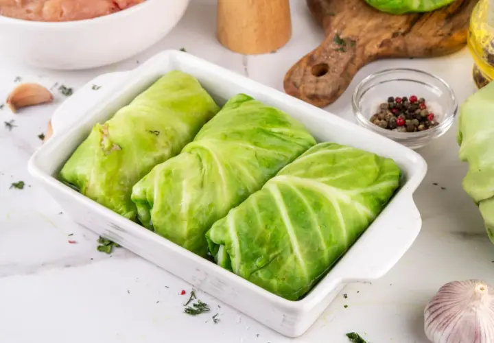 Gotowe do pieczenia gołąbki z młodej kapusty w naczyniu żaroodpornym, obok składniki: mięso mielone, czosnek, przyprawy.