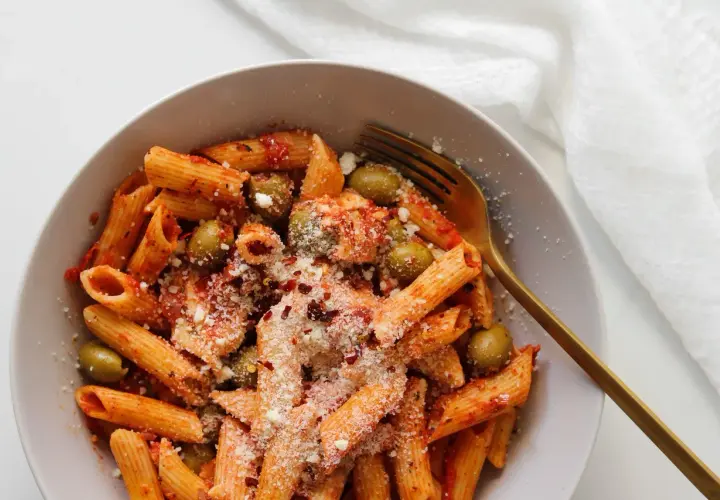 Penne z sosem pomidorowym, oliwkami i startą mozzarellą, posypane płatkami chili. Złoty widelec czeka na pierwszy kęs.