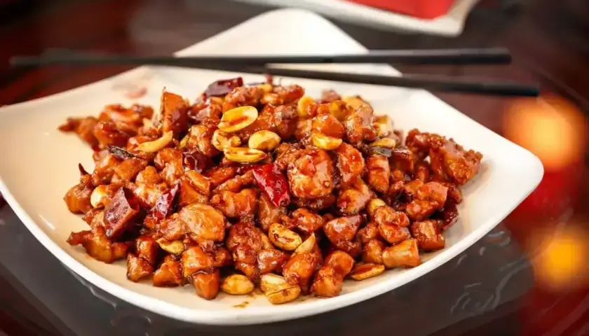 Kurczak King Kong, danie z kawałkami kurczaka w sosie, orzeszkami ziemnymi i papryczkami chili, podane na białym talerzu z pałeczkami.