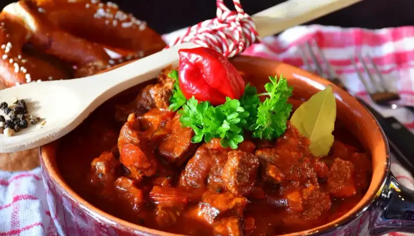 Pyszny gulasz wołowy w glinianej misie, udekorowany papryczką chili i natką pietruszki. Obok leży precel i drewniana łyżka z pieprzem. Idealny przepis na gulasz!