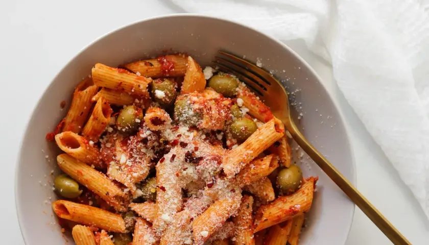Penne z sosem pomidorowym, oliwkami i startą mozzarellą, posypane płatkami chili. Złoty widelec czeka na pierwszy kęs.