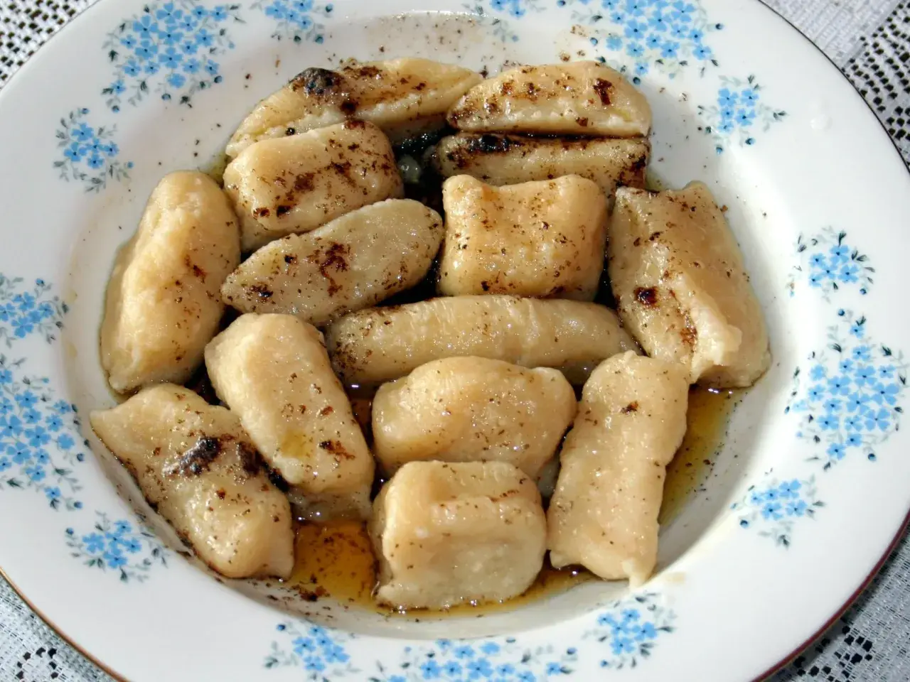 Gotowe pierogi leniwe, polane masłem i posypane cynamonem, czekają na talerzu z niebieskimi kwiatuszkami.