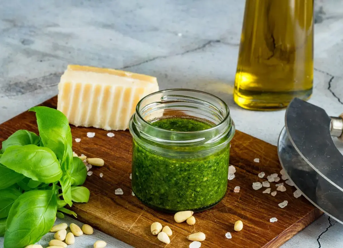Słoiczek domowego pesto z bazylii, idealny sposób, jak zrobić pesto z bazylii na zimę. Obok świeża bazylia, orzeszki piniowe, parmezan i oliwa.