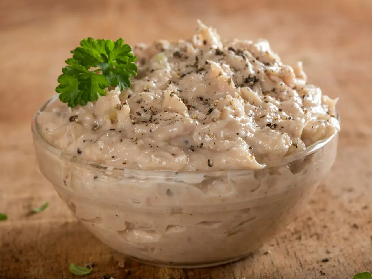 Pyszna pasta z tuńczyka, doprawiona pieprzem i udekorowana natką pietruszki, w szklanej miseczce na drewnianym tle.