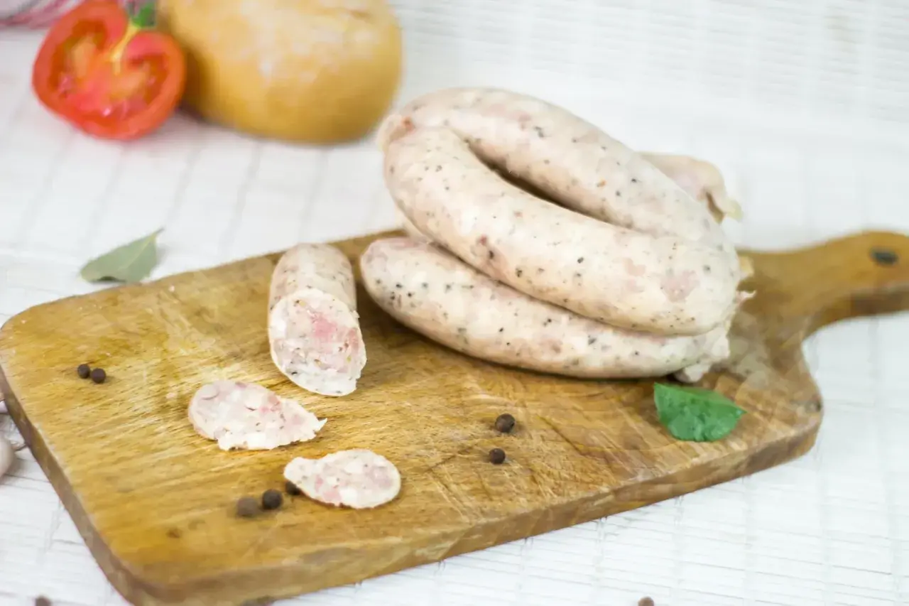Biała kiełbasa na desce, obok pomidor, czosnek i przyprawy. Zastanawiasz się, ile czasu moczymy jelita do kiełbasy?