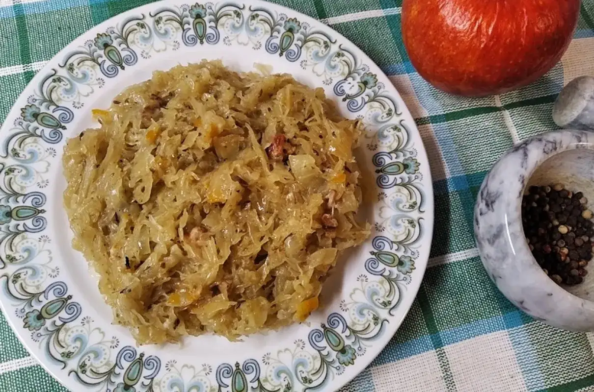 Pyszna kapusta kiszona gotowana, podana na talerzu z ozdobnym wzorem. Obok jabłko i moździerz z pieprzem.