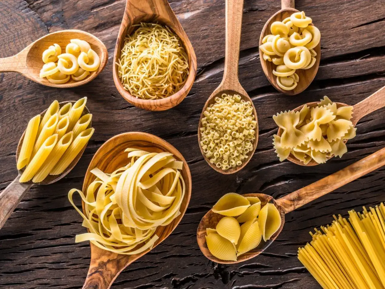 Drewniane łyżki pełne różnorodnych zup z makaronem: penne, farfalle, muszelki, tagliatelle i drobne nitki.