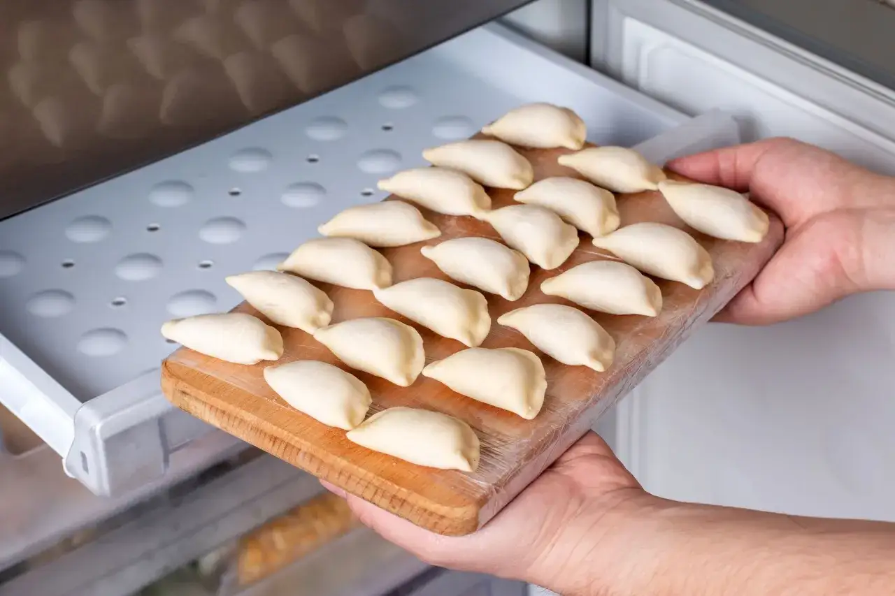 Ręce wkładają deskę z surowymi pierogami do szuflady lodówki. To sposób, jak przechowywać surowe pierogi w lodówce, by zachowały świeżość.