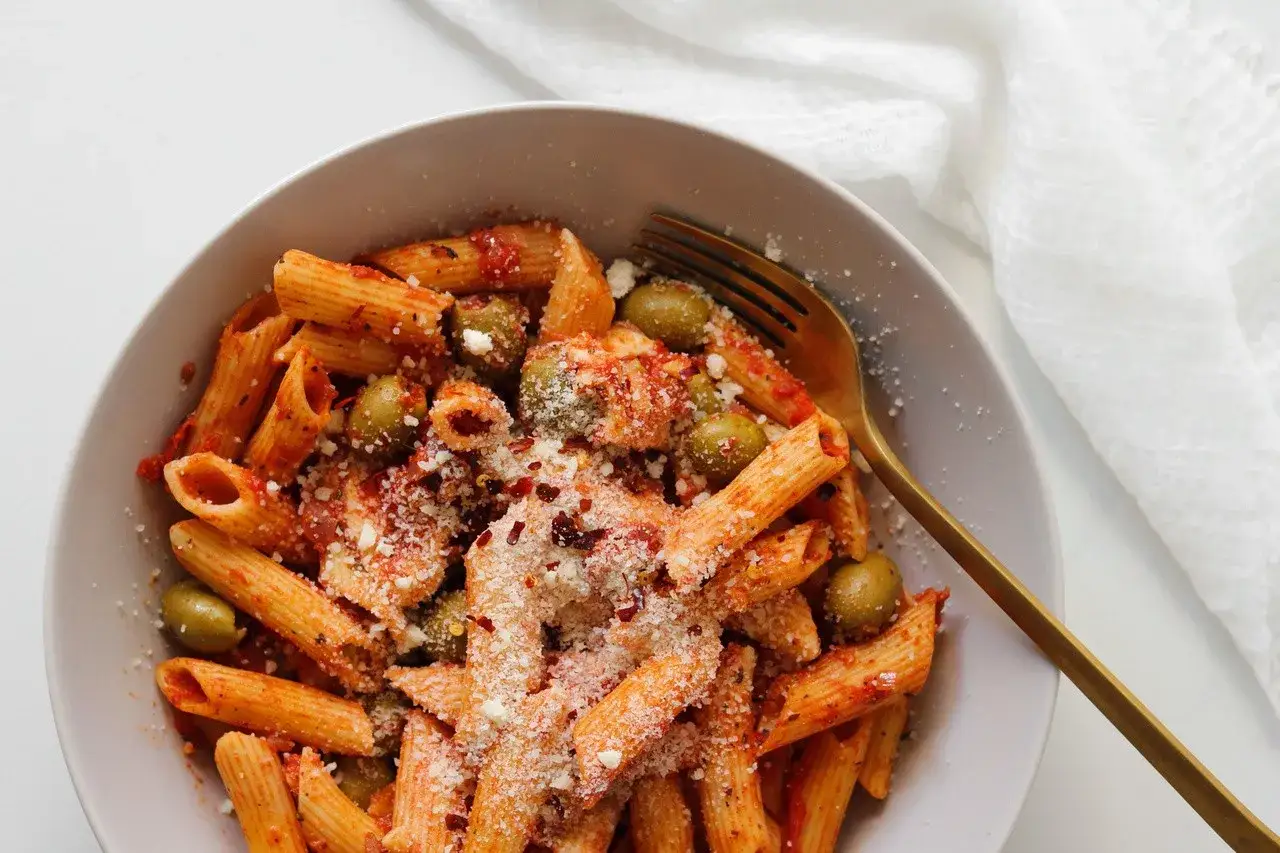 Penne z sosem pomidorowym, oliwkami i startą mozzarellą, posypane płatkami chili. Złoty widelec czeka na pierwszy kęs.