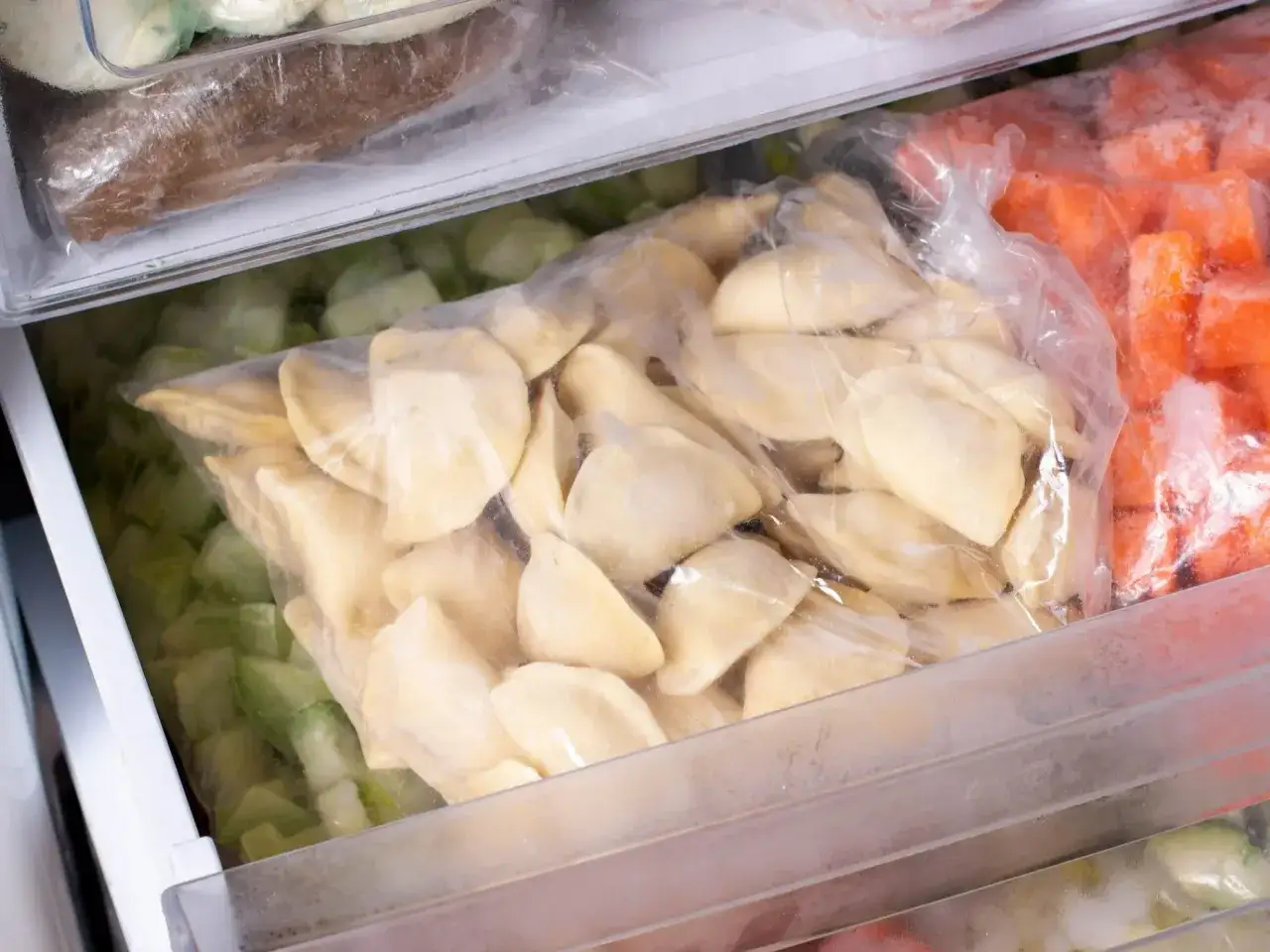 Zamrożone pierogi w woreczku, obok pokrojona marchewka i zielone warzywa. To dobry sposób na to, jak pakować pierogi próżniowo, by zachowały świeżość.