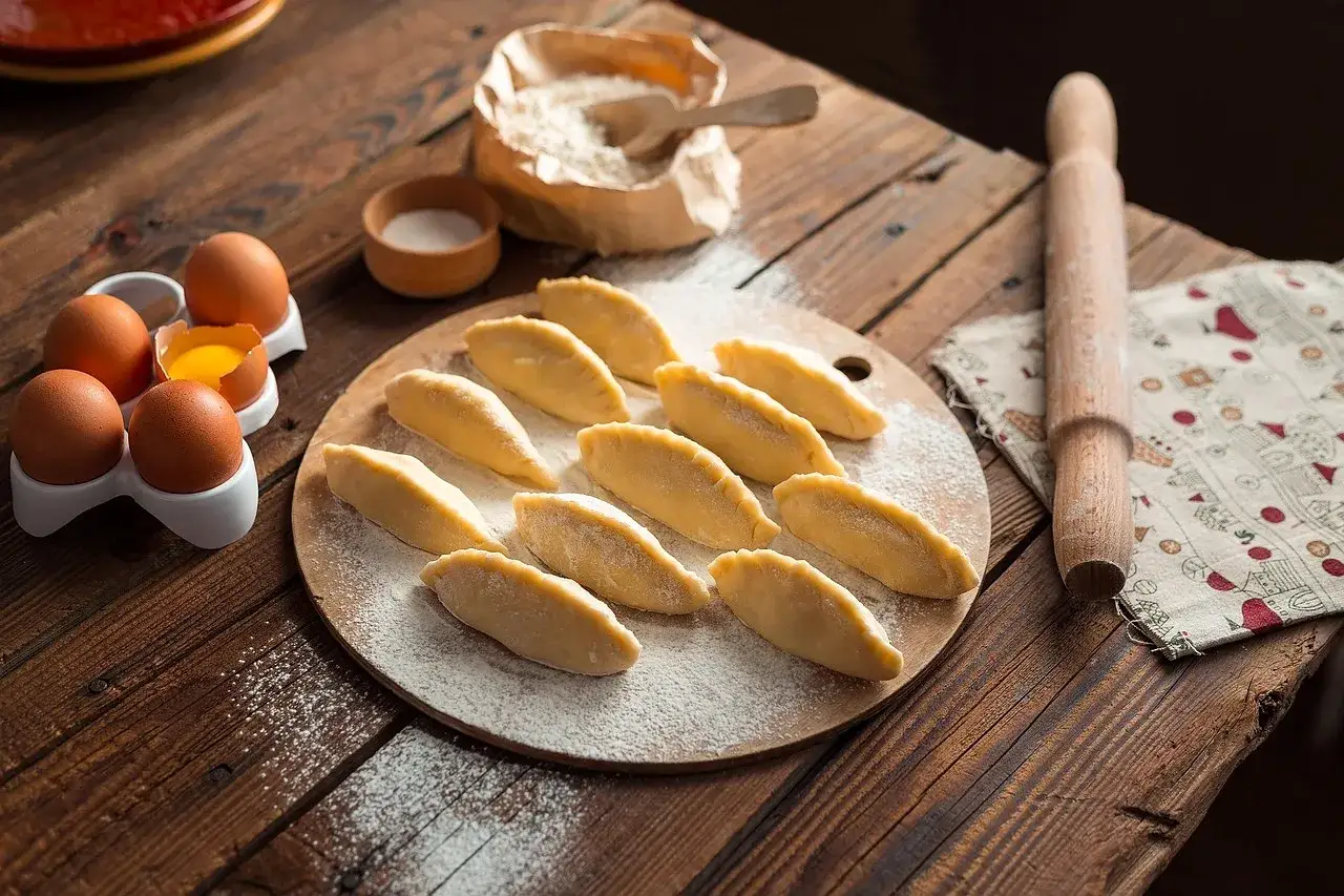 Surowe pierogi na desce, jajka, mąka i wałek. Czy ciasto na pierogi można zamrozić? Tak, można je zamrozić.