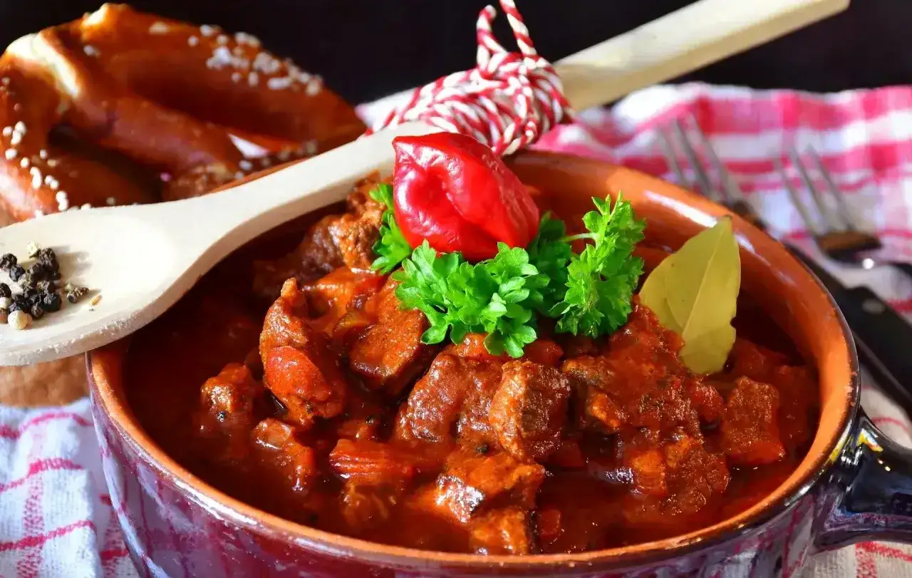 Pyszny gulasz wołowy w glinianej misie, udekorowany papryczką chili i natką pietruszki. Obok precel i drewniana łyżka z pieprzem. Idealny przepis na gulasz.