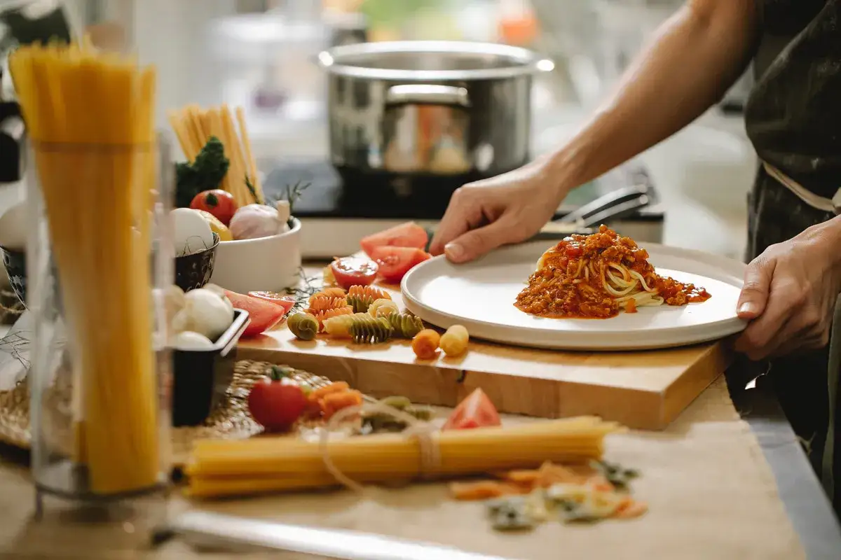 Gotowanie spaghetti bolognese to coś, co każdy potrafi ugotować. Na talerzu porcja makaronu z sosem, obok składniki: pomidory, czosnek, r&oacute;żne rodzaje makaronu.