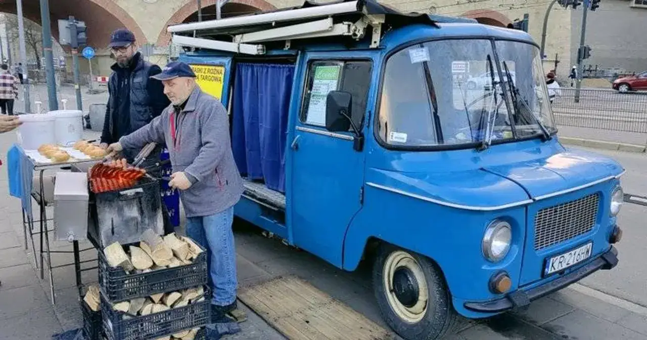 Sprzedawca przygotowuje kiełbaski z niebieskiej Nyski. Obok stoją bułki i drewno do grillowania.