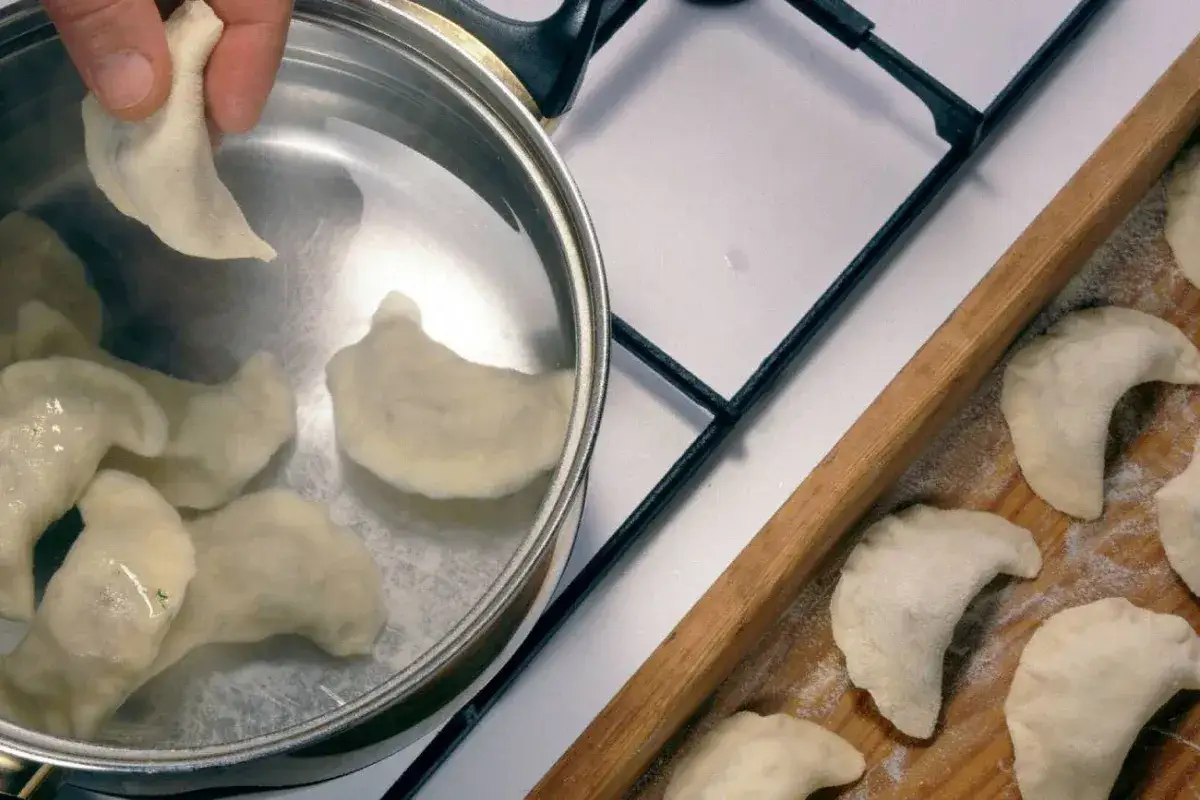 Gotowanie pierogów. Czy płukać pierogi po ugotowaniu? Na desce leżą surowe pierogi, a w garnku gotują się już pierwsze.