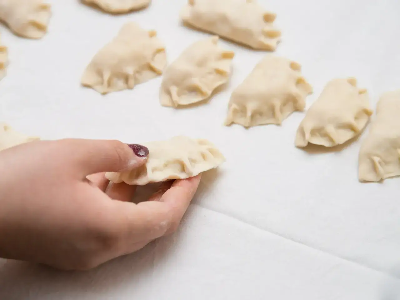 Dłoń trzyma surowe pierogi, pokazując, jak zrobić pierogi. W tle widać więcej gotowych do gotowania pierogów.