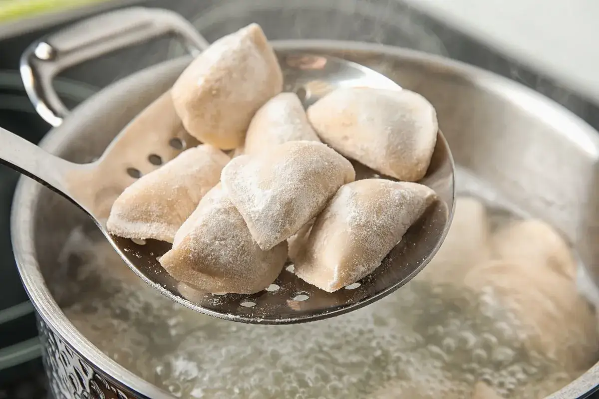 Pierogi wrzuca się do gotującej wody. Łyżka cedzakowa z surowymi pierogami nad garnkiem z bulgoczącą wodą.